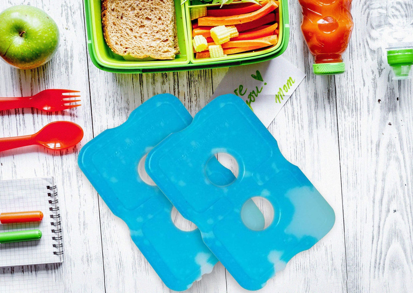 Healthy Packers blue ice packs displayed on a white wooden table next to a green bento box filled with sandwiches, vegetables, and an apple