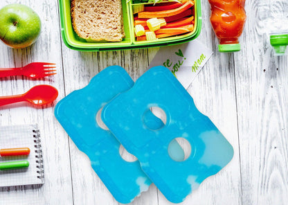 Healthy Packers blue ice packs displayed on a white wooden table next to a green bento box filled with sandwiches, vegetables, and an apple