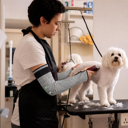 Woman wearing grey Schwer ANSI A6 cut-resistant sleeves and apron while grooming a white dog on a trimming table in a pet salon.