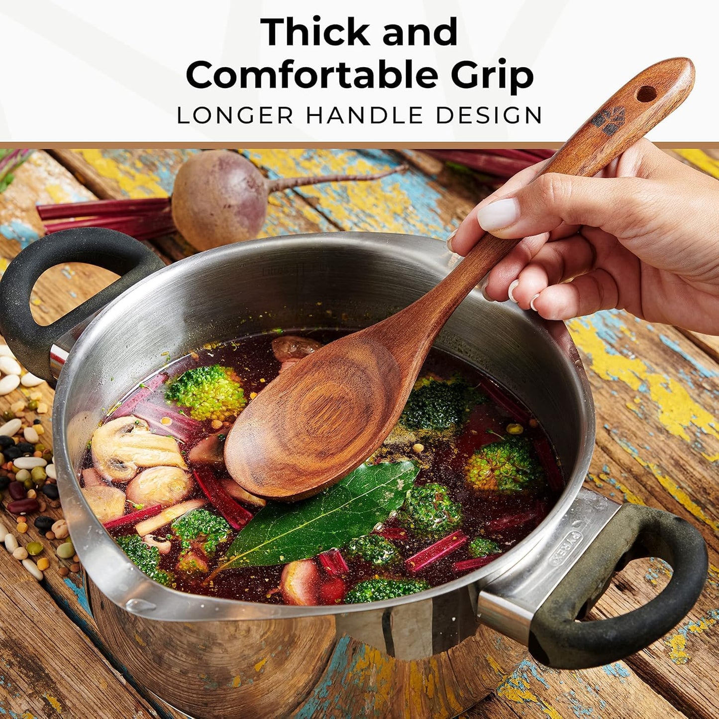 Close-up of a hand holding a long-handled Woodenhouse teak wood spoon lifting vegetables from a full soup pot, with 'Thick and Comfortable Grip' text visible.