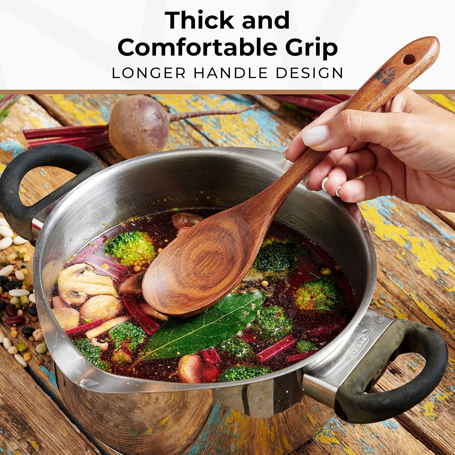 Close-up of a hand holding a long-handled Woodenhouse teak wood spoon lifting vegetables from a full soup pot, with 'Thick and Comfortable Grip' text visible.